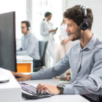 Homem sentado na frente de um computador fala com alguém pelo headset